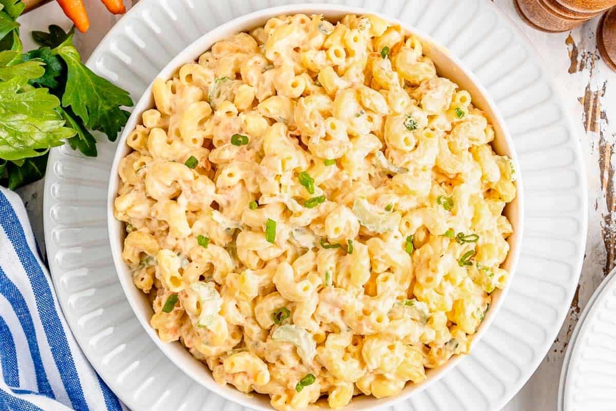 A white bowl filled with creamy macaroni salad garnished with chopped green herbs, placed on a white plate.