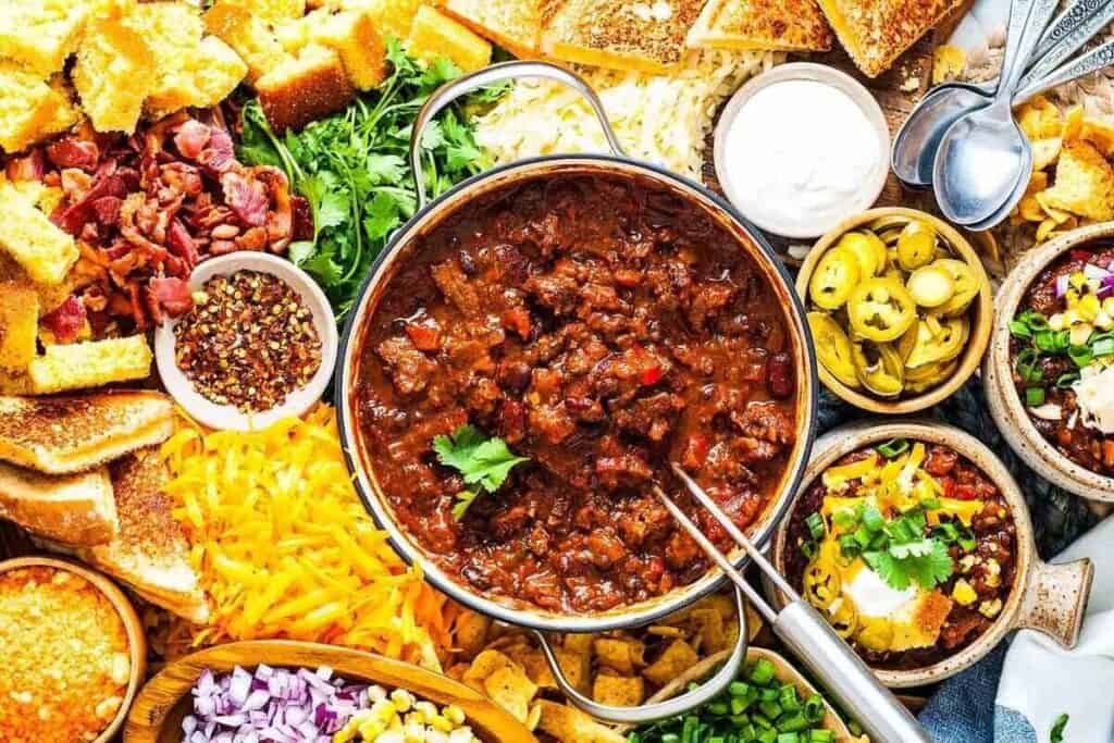 A pot of chili surrounded by toppings including shredded cheese, jalape&ntilde;os, sour cream, chopped onions, cornbread, bacon bits, and cilantro.