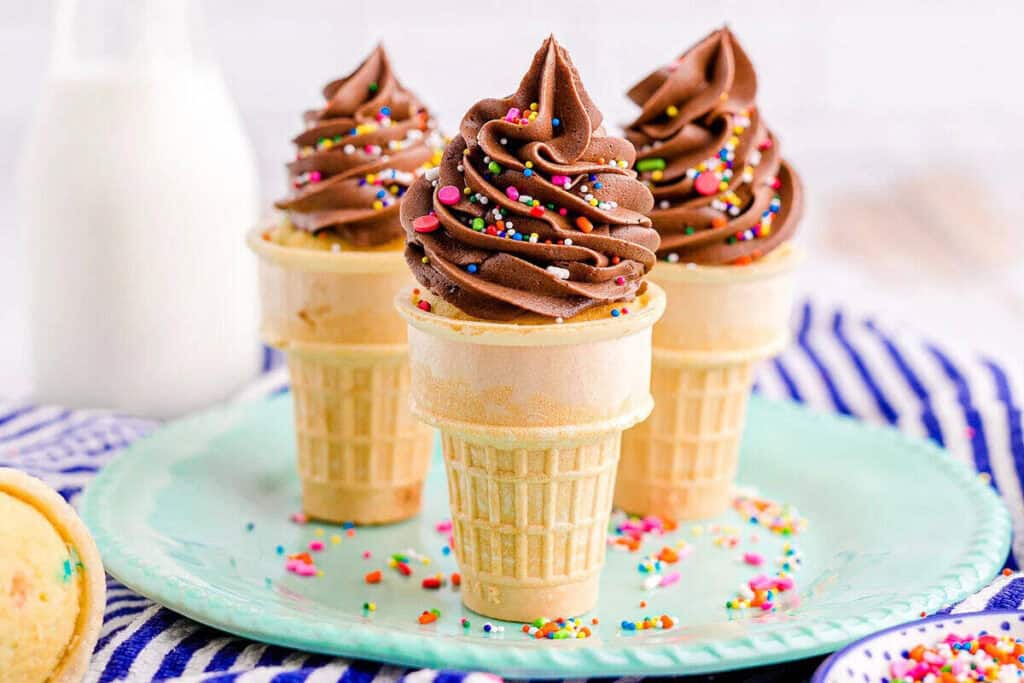 Three ice cream cone cupcakes with chocolate frosting and colorful sprinkles are arranged on a mint green plate, with a milk bottle and striped cloth in the background.