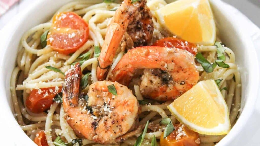 A bowl of spaghetti topped with grilled shrimp, cherry tomatoes, lemon wedges, grated cheese, and chopped fresh herbs.