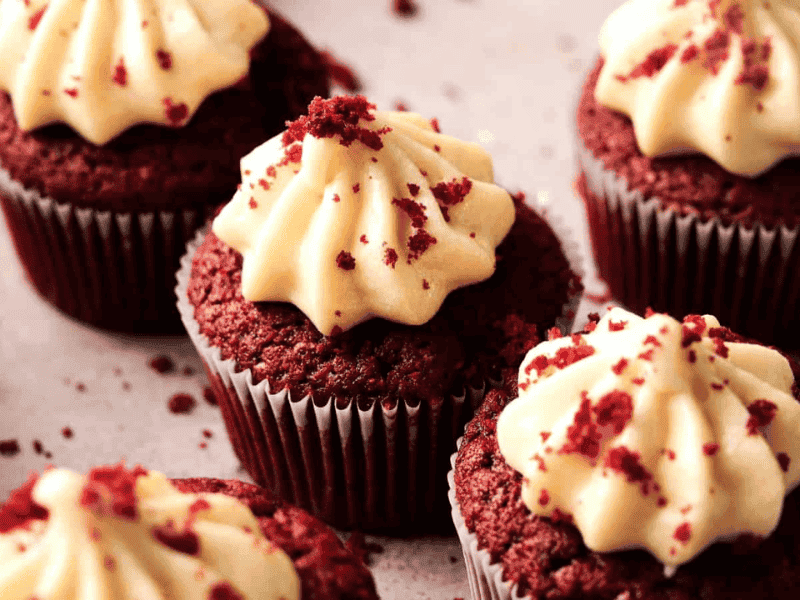 Four red velvet cupcakes with cream cheese frosting are arranged on a light surface, with red cake crumbs sprinkled on top and around them.