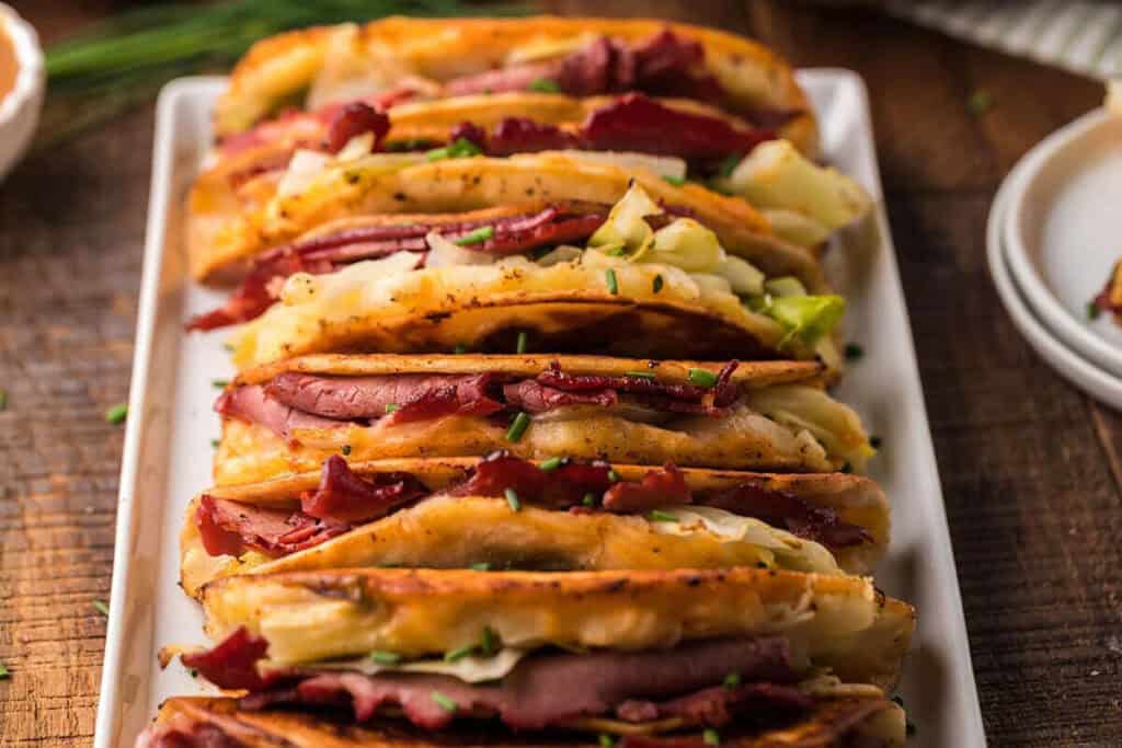 A row of crispy sandwiches filled with sliced meat and cabbage, garnished with chopped chives, served on a rectangular white plate.