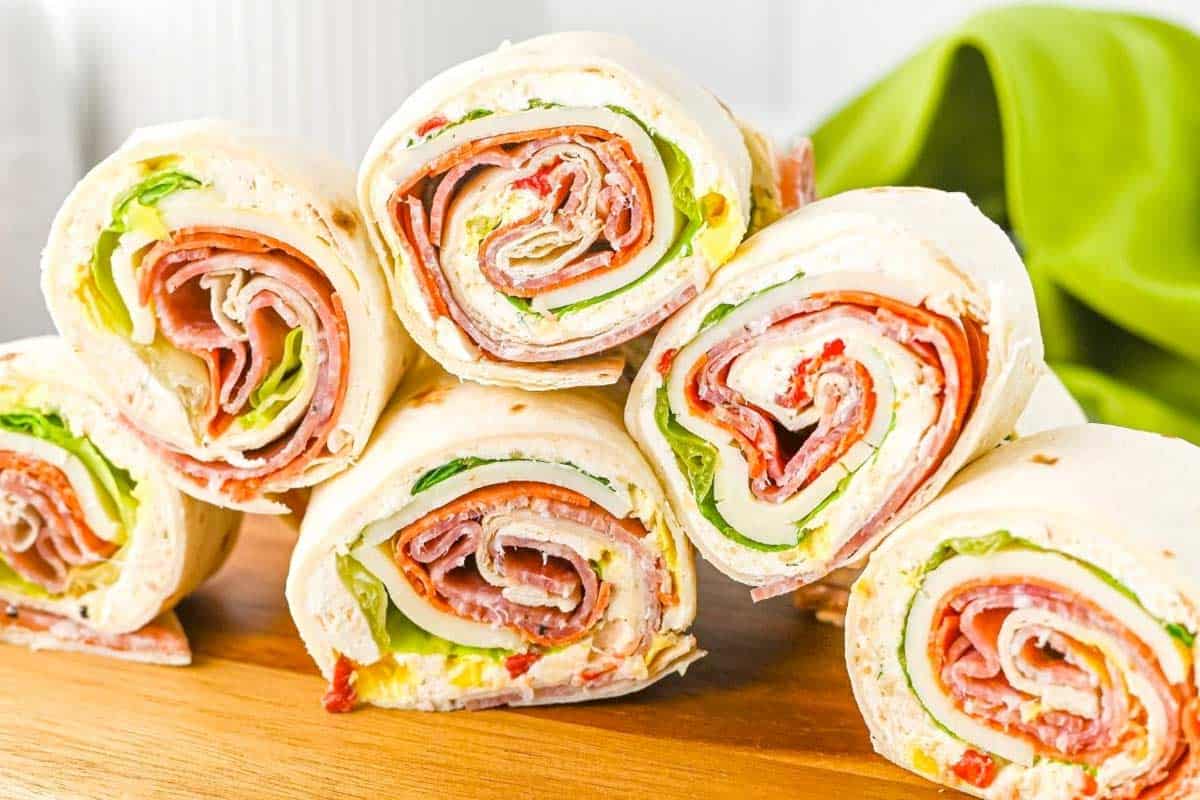 Italian Pin Wheels are stacked on a wooden surface, revealing layers of deli meat, cheese, lettuce, and other fresh vegetables.