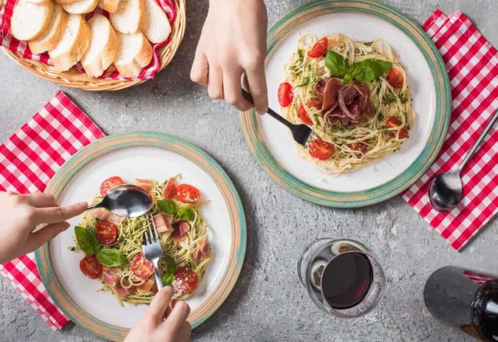 Two plates of pasta with tomatoes and greens, a basket of sliced bread, and a glass of red wine on a gray table with red checkered napkins capture the inviting atmosphere of a real Italian restaurant; hands holding utensils.