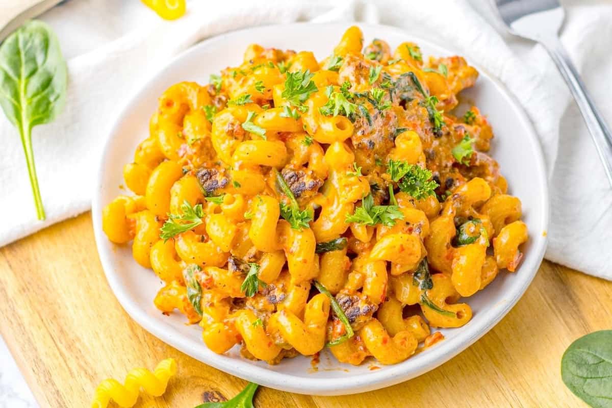 A plate of creamy macaroni pasta mixed with vegetables and garnished with chopped parsley, placed on a wooden surface.
