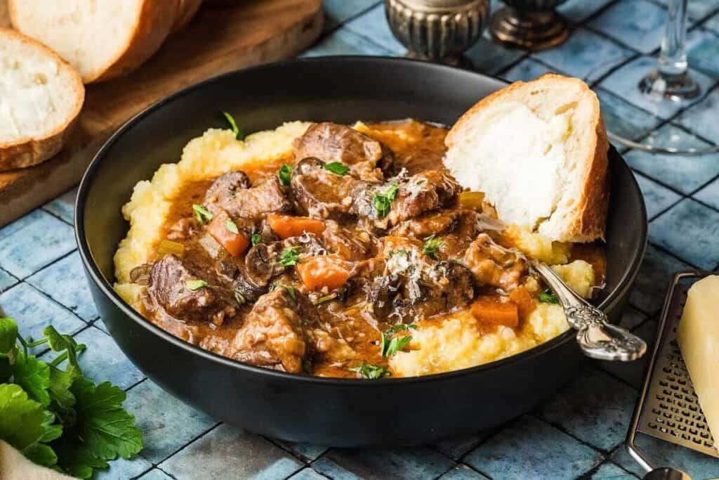 A bowl of beef stew with carrots served over creamy polenta, garnished with herbs, with a slice of bread and a spoon on the side.