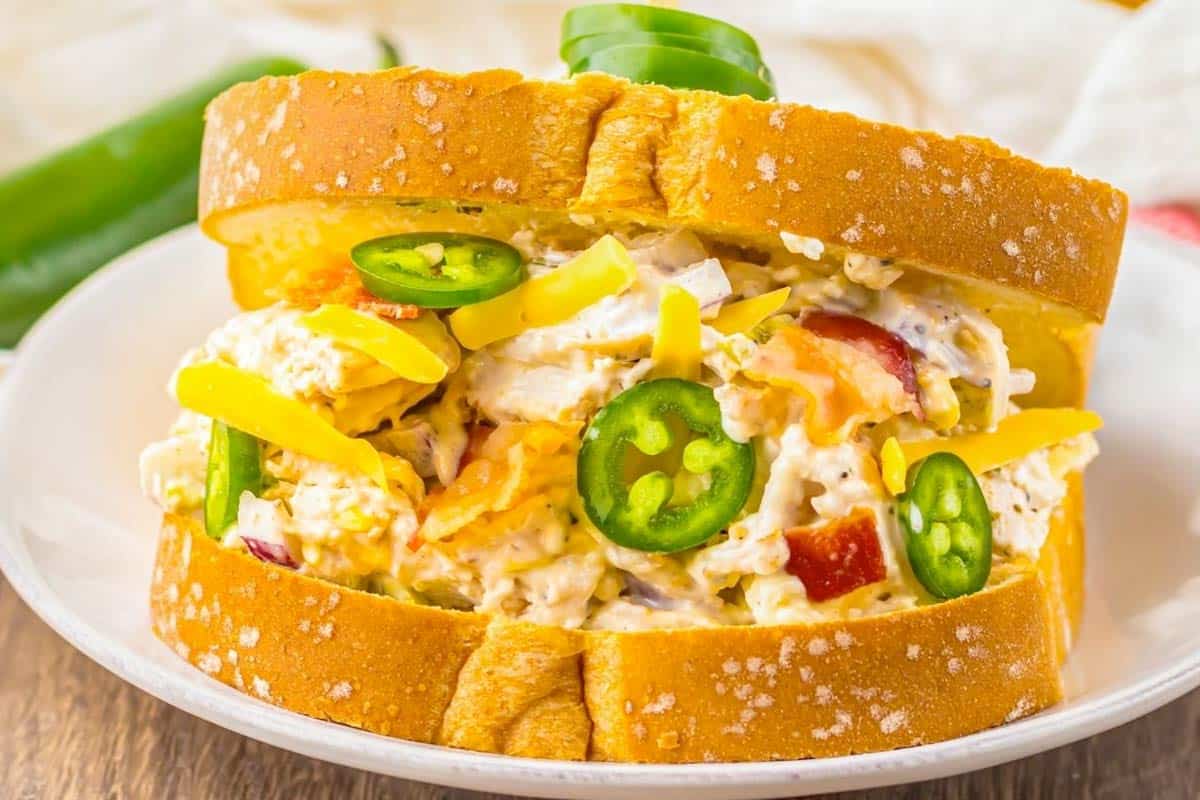 A Jalapeno Popper Chicken Salad sandwich with shredded cheese, bacon, and sliced jalape&ntilde;os on thick white bread sits on a white plate.