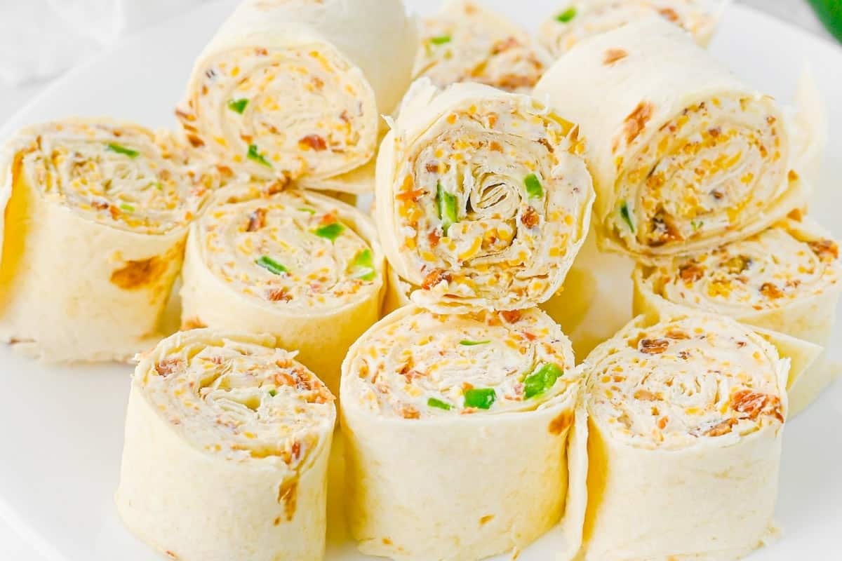 Close-up of sliced tortilla pinwheels filled with a creamy mixture containing shredded cheese, green bell pepper pieces, and bits of bacon. The pinwheels are arranged in a stack.