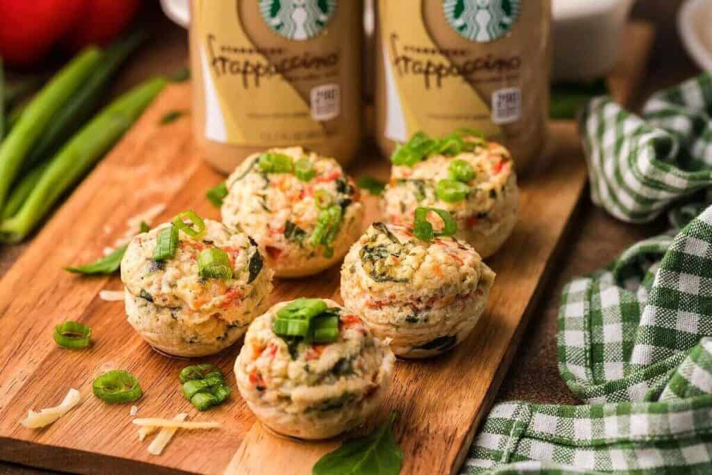 Five egg bites with vegetables and green onions are arranged on a wooden board, inspired by Starbucks Copy Cat Recipes, with Frappuccino bottles in the background and a green checkered cloth to the side.