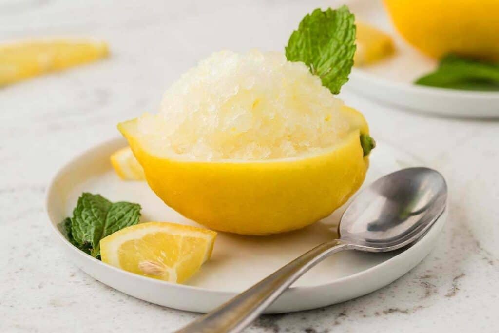 Shaved ice served in a hollowed lemon half, garnished with a mint leaf and lemon wedge, on a white plate with a spoon&mdash;a refreshing no bake treat perfect for warm days.