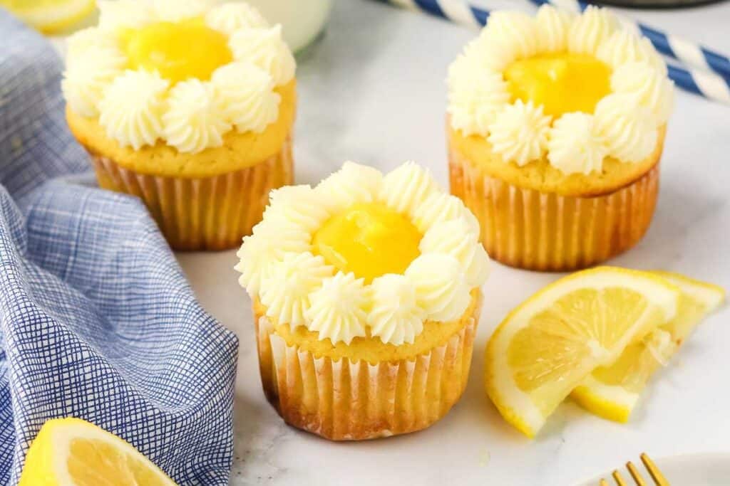 Three lemon cupcakes with white frosting and a lemon curd center are arranged on a marble surface, next to a blue checkered cloth and lemon slices.