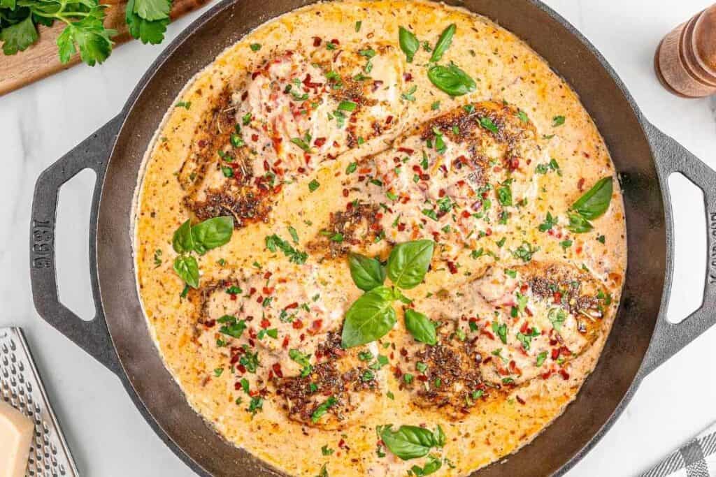 Four seasoned chicken breasts simmer in a creamy sauce, garnished with chopped herbs and basil leaves, in a black cast iron skillet.