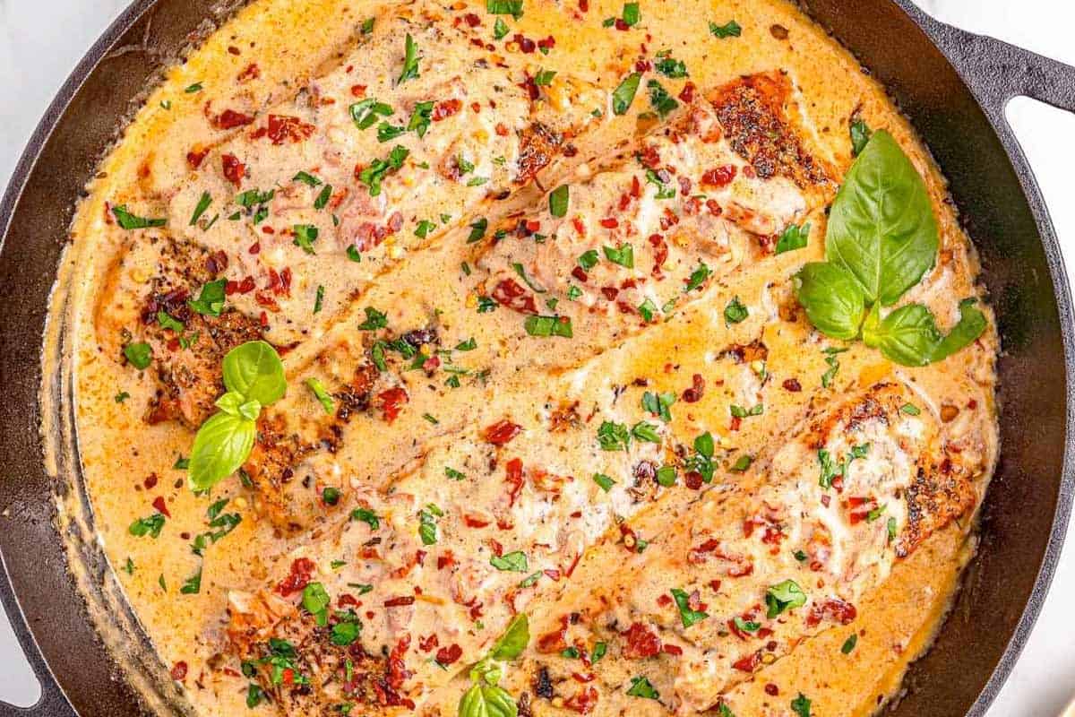 Four salmon fillets in a creamy sauce with chopped herbs, sun-dried tomatoes, and basil leaves, cooked in a black skillet.