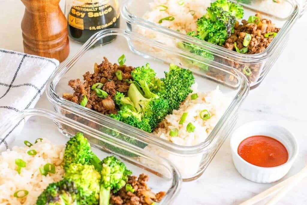 Three glass meal prep containers with ground beef, steamed broccoli, and white rice, garnished with green onions, on a white surface with soy sauce and sriracha nearby.