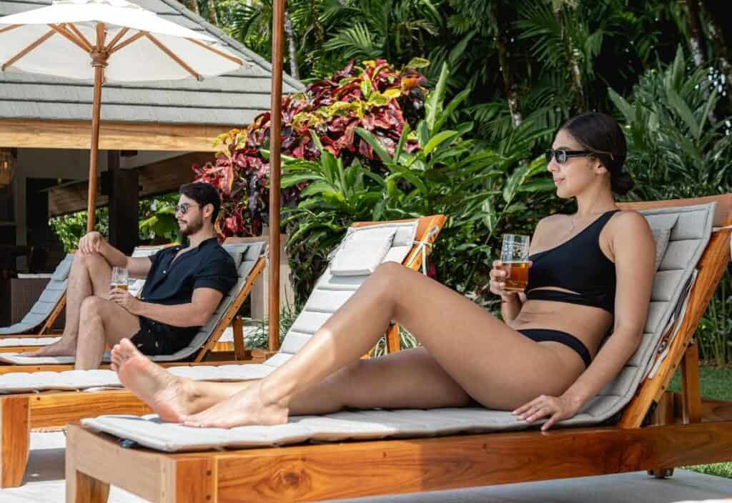 Two people in swimwear relax on lounge chairs by a pool, surrounded by greenery; both wear sunglasses while enjoying drinks—capturing the perfect vibe for a spring break or March travel getaway.