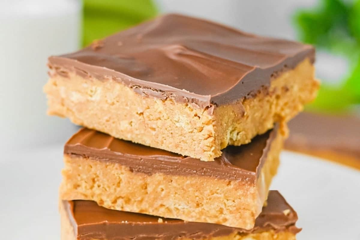 Three stacked peanut butter bars with a thick chocolate layer on top, displayed on a white plate with a blurred green background.