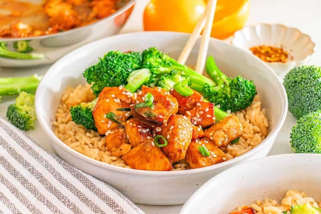 A bowl of brown rice topped with glazed chicken pieces, steamed broccoli, sesame seeds, and chopped green onions—an inviting take on Asian-inspired dishes—with chopsticks resting on the side.