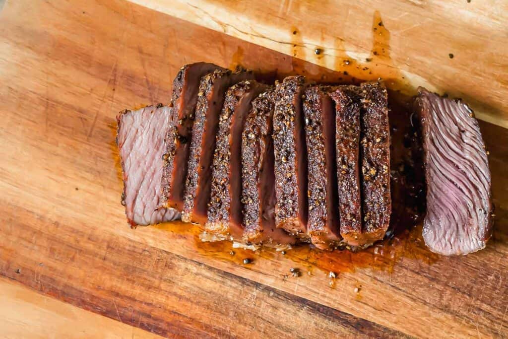 Sliced roast beef with a peppered crust arranged on a wooden cutting board.