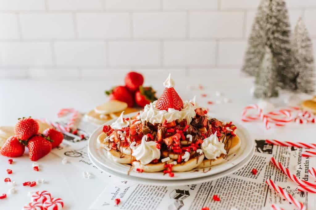 A plate of waffles topped with whipped cream, chopped strawberries, nuts, and chocolate, surrounded by strawberries, candy canes, and Christmas decorations&mdash;perfect as delightful Valentines treats on a festive white surface.