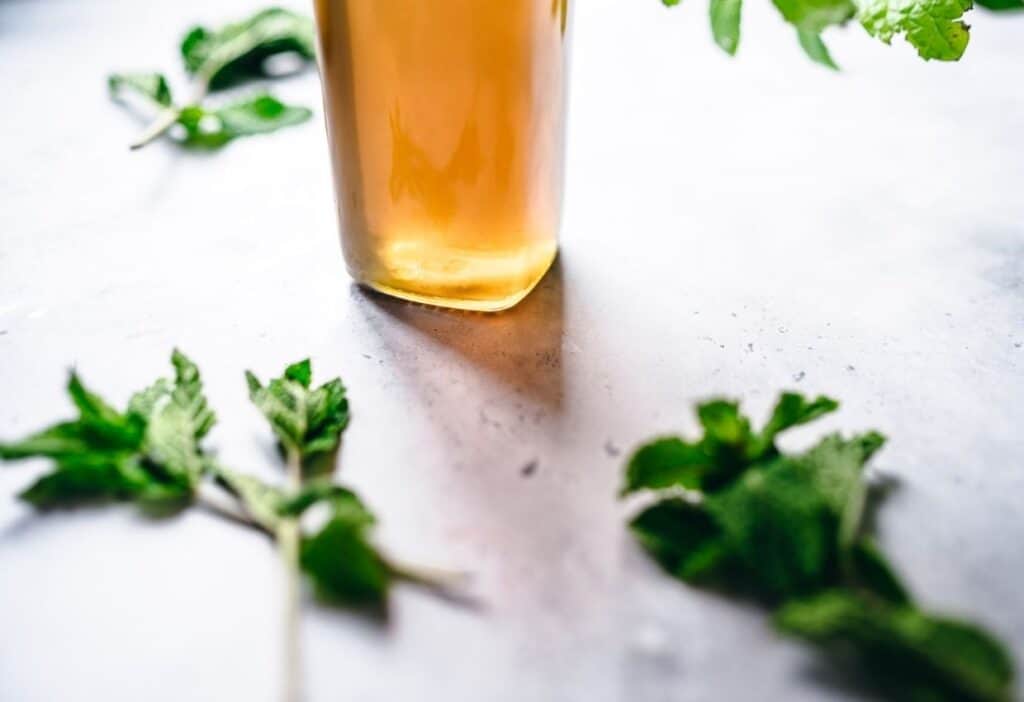 A glass of amber-colored liquid sits on a light surface, surrounded by fresh green mint leaves.
