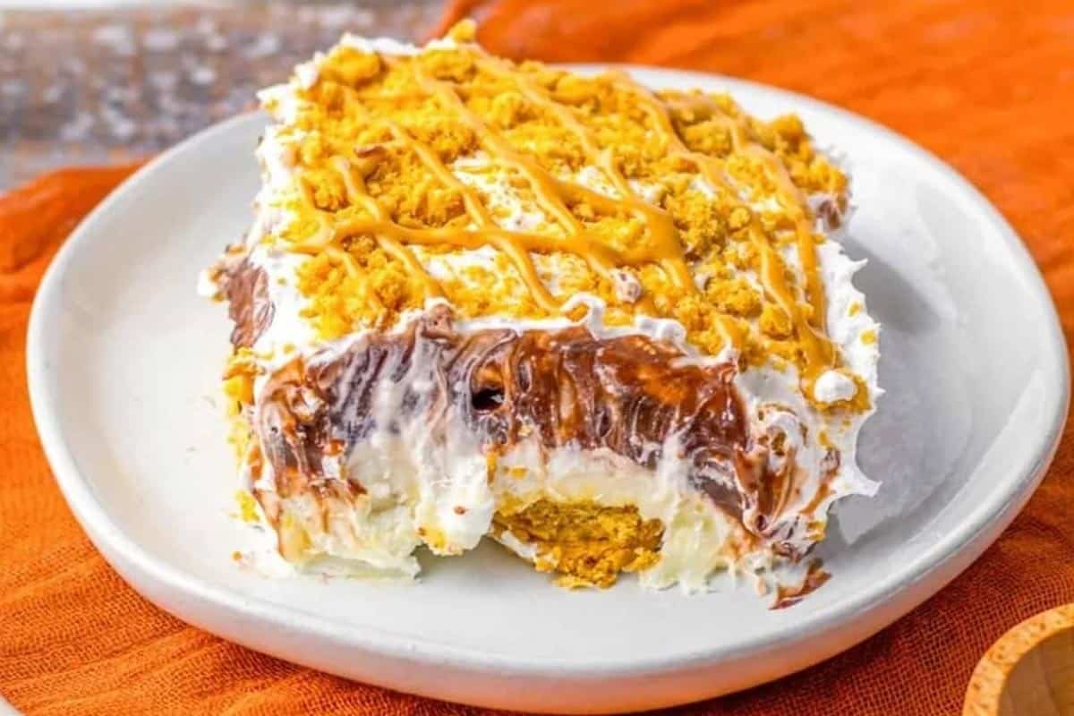 A slice of layered dessert with whipped topping, chocolate, and crumbly crust, drizzled with caramel, served on a white plate with an orange cloth underneath.