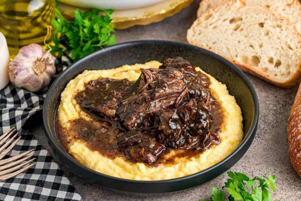 A bowl of creamy polenta topped with braised beef in a dark sauce, surrounded by bread, garlic, parsley, and a checkered napkin&mdash;a comforting favorite among tasty beef recipes.