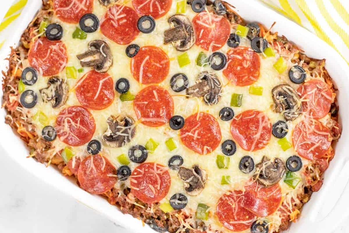 A baked casserole topped with melted cheese, pepperoni slices, black olives, mushrooms, and diced green peppers in a white dish.