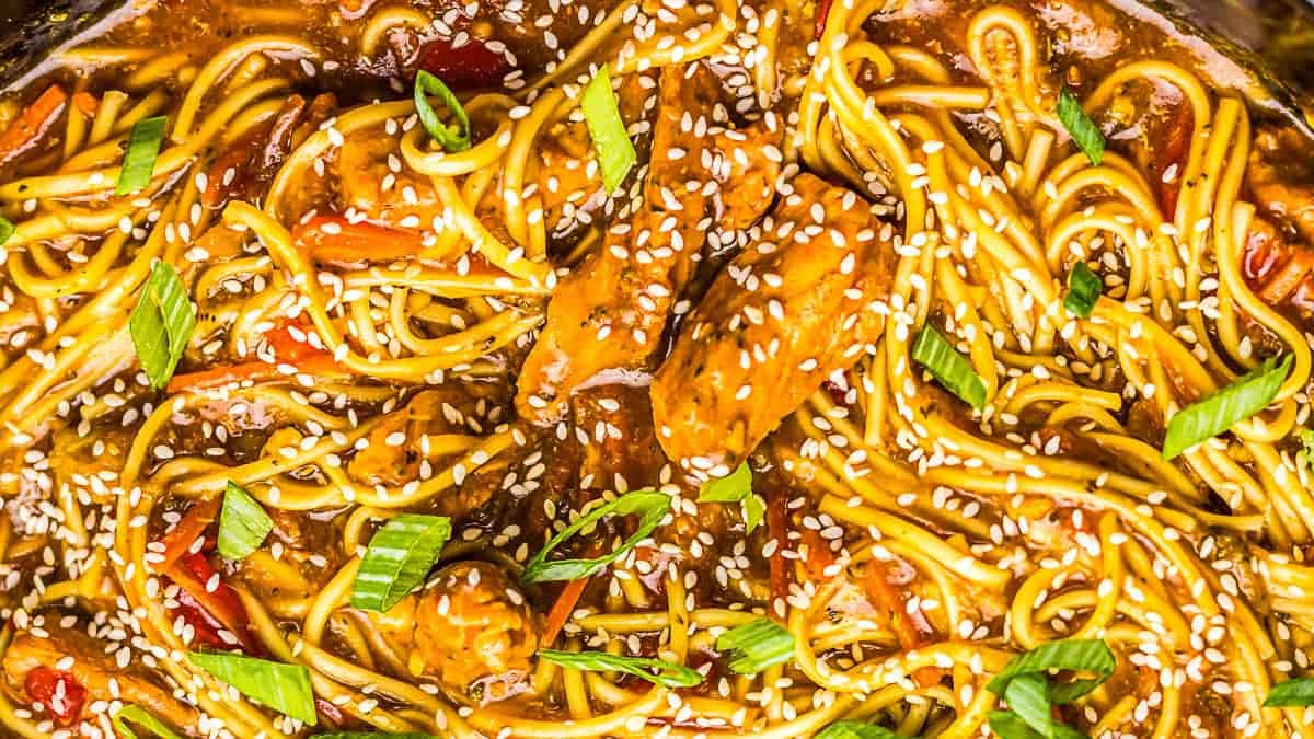 A close-up of cooked noodles with chicken, sliced green onions, red peppers, sesame seeds, and a brown sauce.