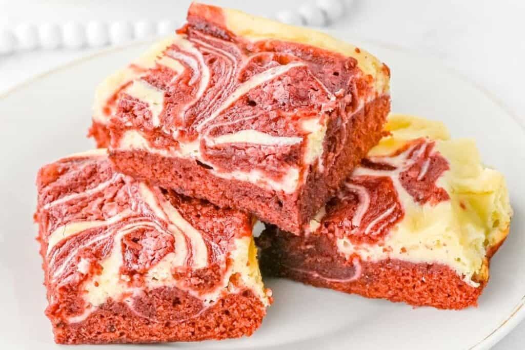 Three square red velvet cheesecake brownies with cream cheese swirls stacked on a white plate.