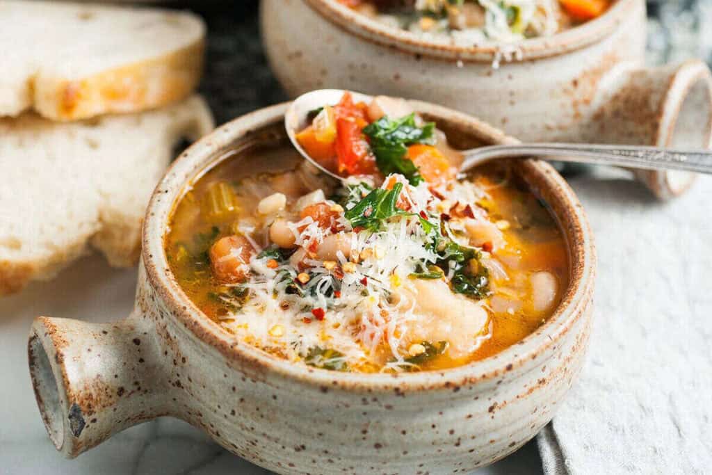 A bowl of vegetable and bean soup topped with grated cheese and herbs, with a spoon resting on the bowl and slices of bread in the background.