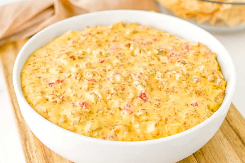 A white bowl filled with creamy cheese dip mixed with ground meat and diced tomatoes, placed on a wooden surface.