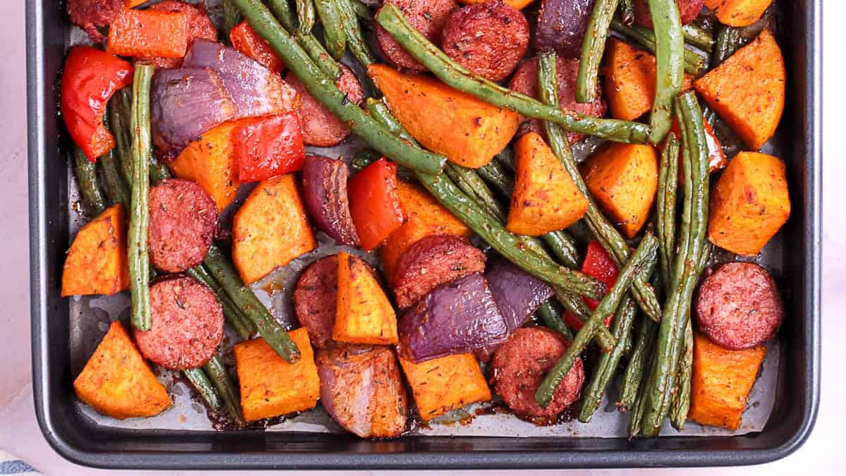 Sheet pan with roasted green beans, sliced sausage, sweet potatoes, red onion, and red bell pepper.