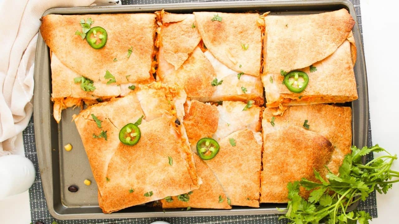 A baking tray with baked, square-cut quesadillas topped with jalape&ntilde;o slices and cilantro, with a bunch of fresh cilantro beside the tray.