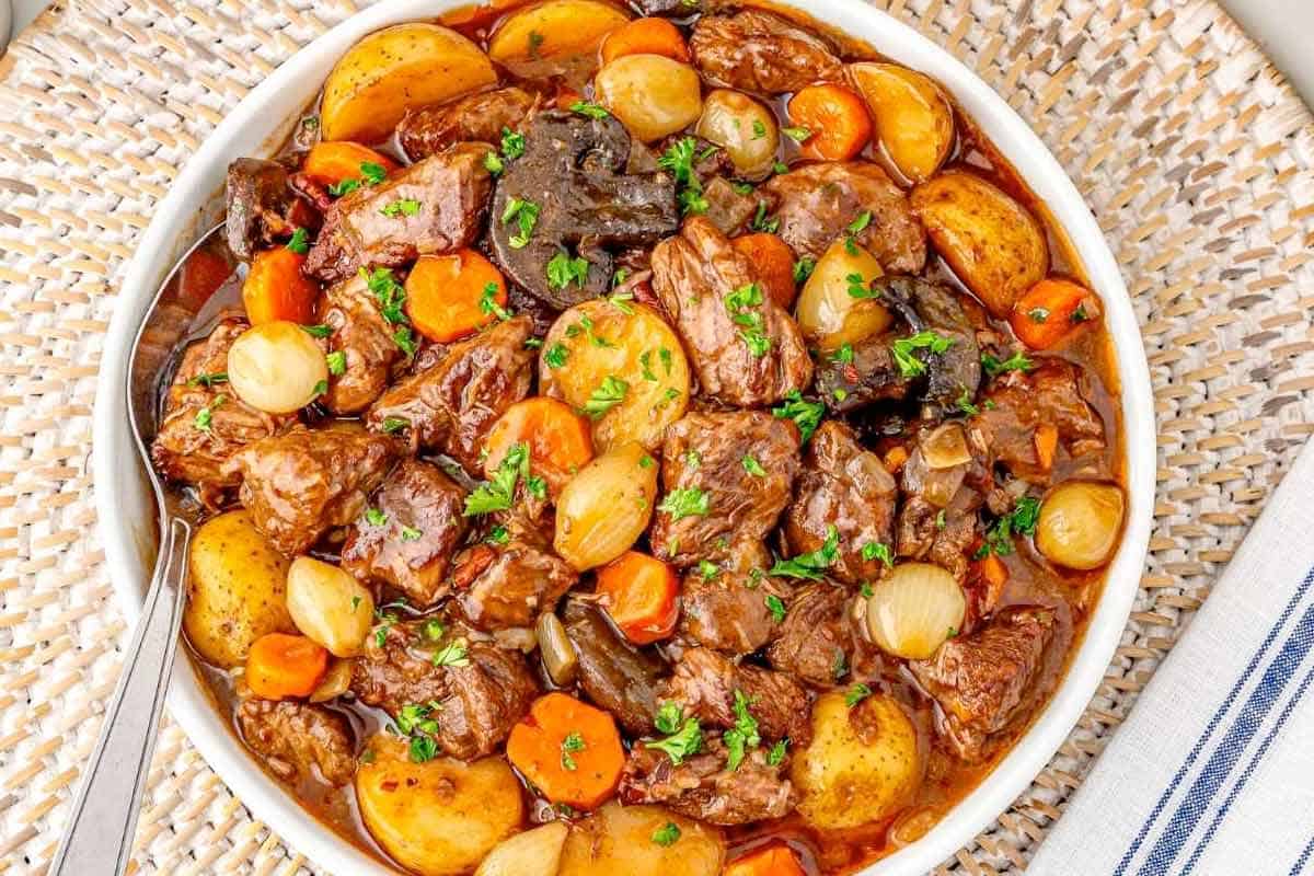 A bowl of beef stew with potatoes, carrots, pearl onions, and mushrooms in a rich brown sauce, garnished with chopped parsley.