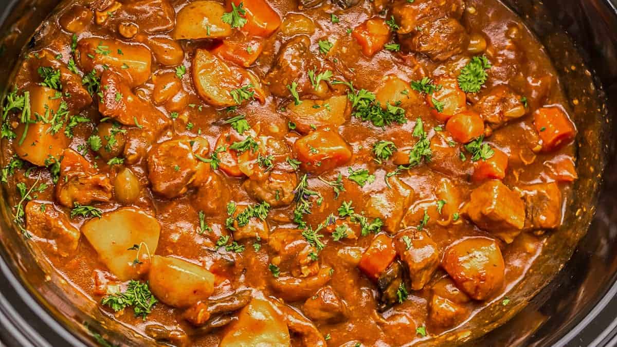 Beef stew with potatoes, carrots, and herbs in a thick brown sauce, garnished with chopped parsley, shown in a cooking pot.