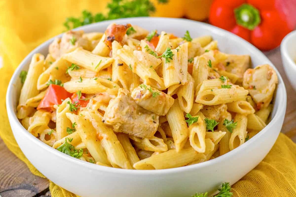 A bowl of penne pasta with pieces of chicken, red bell peppers, and garnished with chopped parsley.