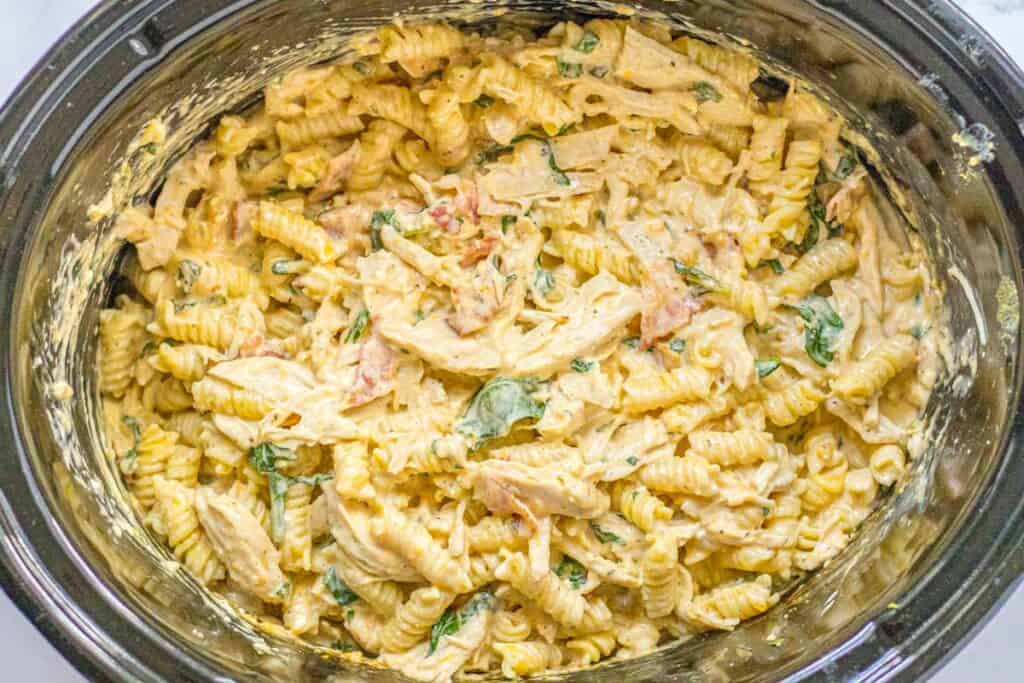 Cooked rotini pasta mixed with shredded chicken, spinach, tomato, and a creamy sauce in a slow cooker.