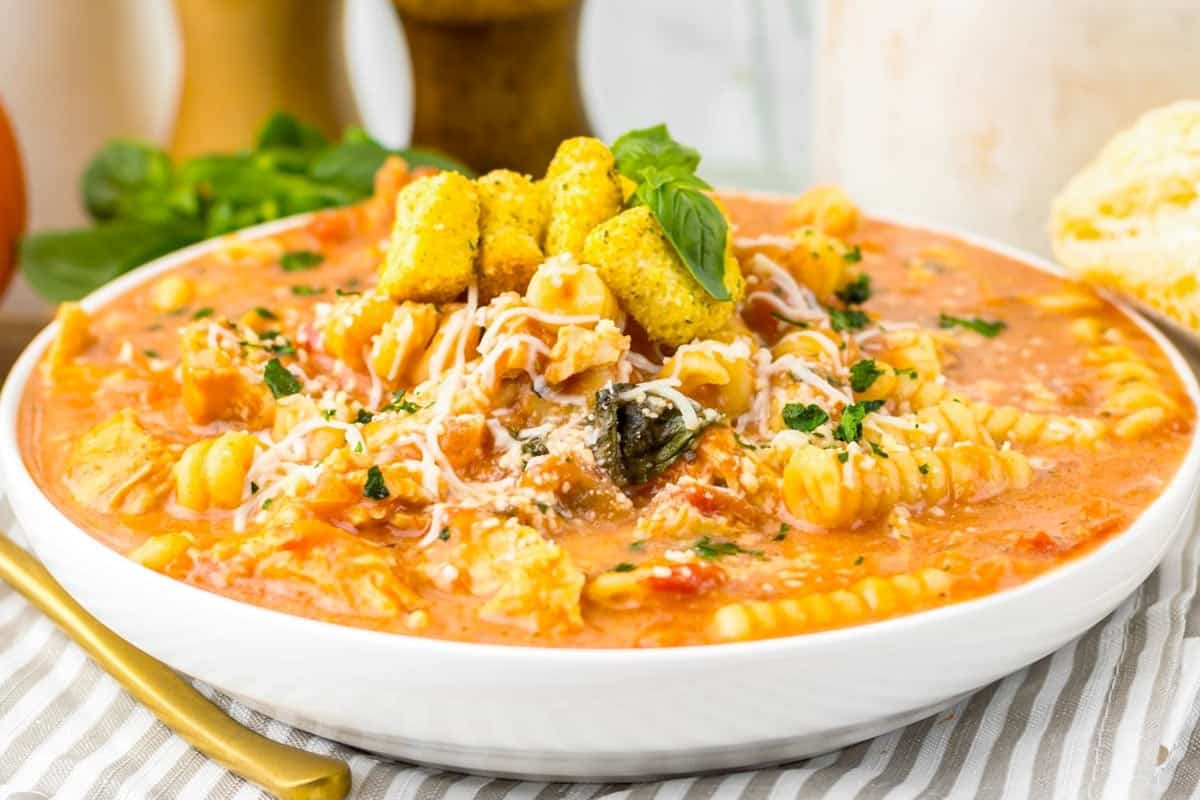 A bowl of creamy tomato pasta soup topped with shredded cheese, croutons, and fresh herbs, with bread on the side.