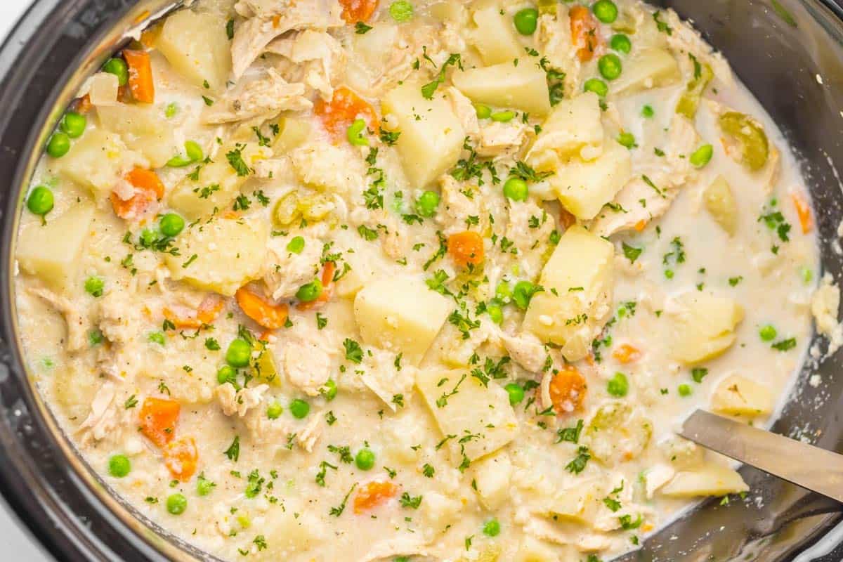 A close-up of a creamy chicken stew with potatoes, carrots, peas, and herbs in a slow cooker, with a metal serving spoon visible.