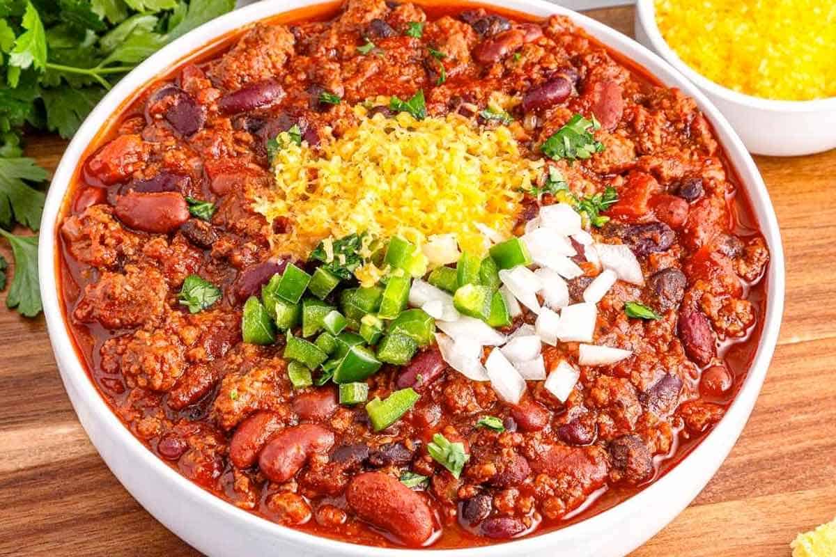 A bowl of chili topped with shredded cheese, chopped onions, and diced jalapeños, with a side dish of shredded cheese in the background.