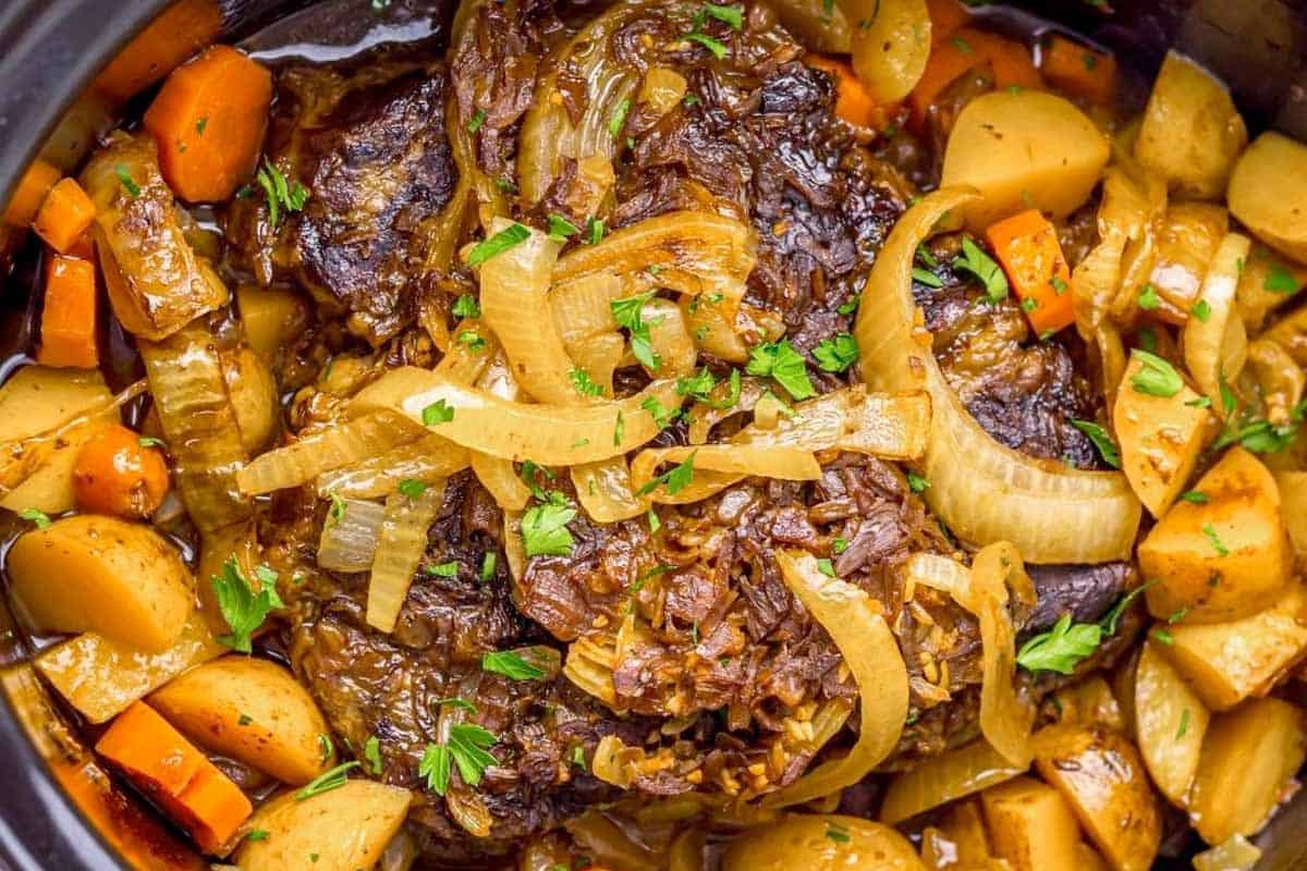 A close-up of a pot roast with sliced onions, carrots, and potatoes, garnished with chopped parsley.