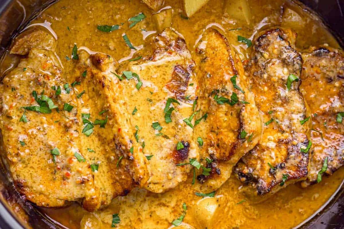 Four pork chops in a creamy, orange-colored sauce, garnished with chopped herbs and served in a pan.