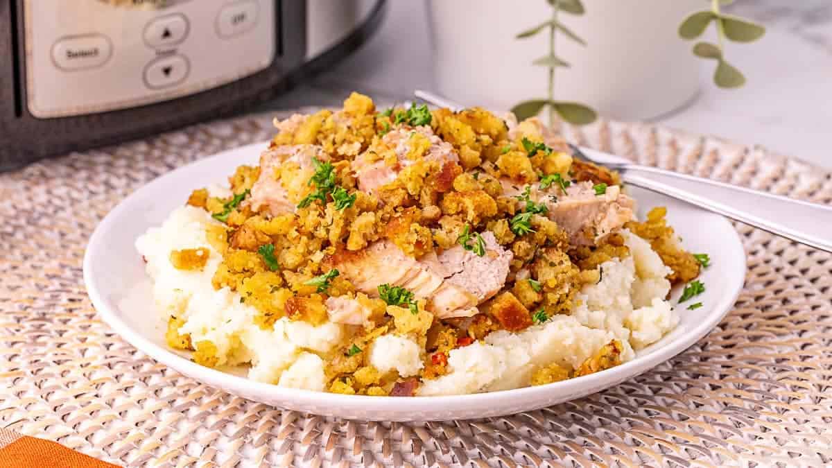 A plate of mashed potatoes topped with sliced turkey and stuffing, garnished with chopped parsley, sits on a woven placemat with a fork beside it.