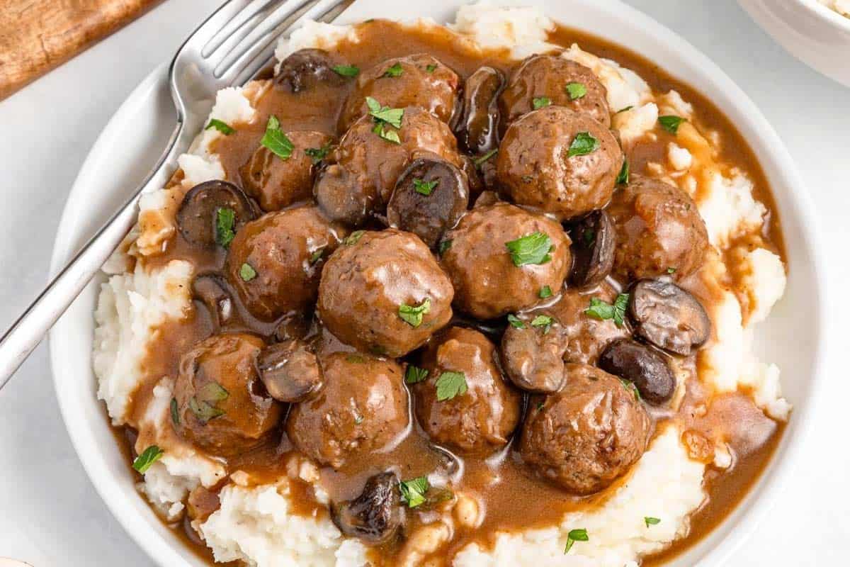 A plate of mashed potatoes topped with meatballs, sliced mushrooms, and brown gravy, garnished with chopped parsley, with a fork on the side.