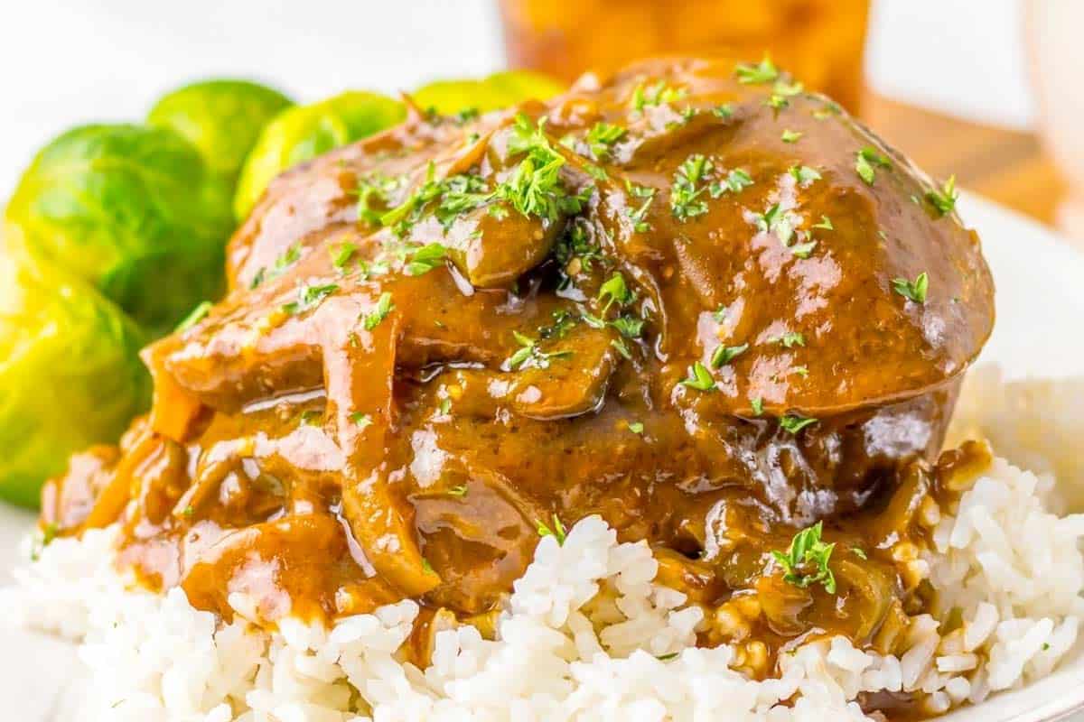 A serving of rice topped with Salisbury steak in brown gravy, garnished with herbs, and accompanied by Brussels sprouts.