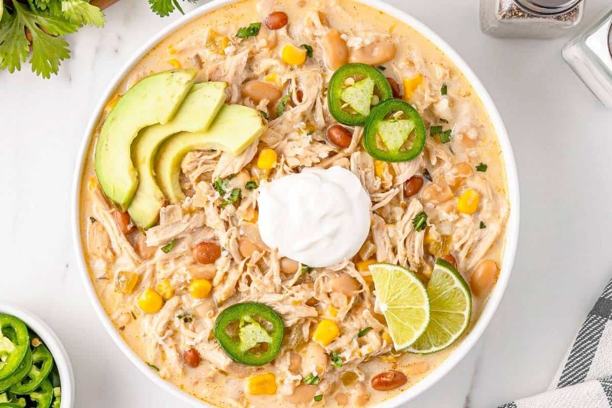 A bowl of creamy White Chicken Chili with shredded chicken, corn, beans, jalape&ntilde;o slices, avocado, lime wedge, and sour cream, garnished with herbs on a marble surface.