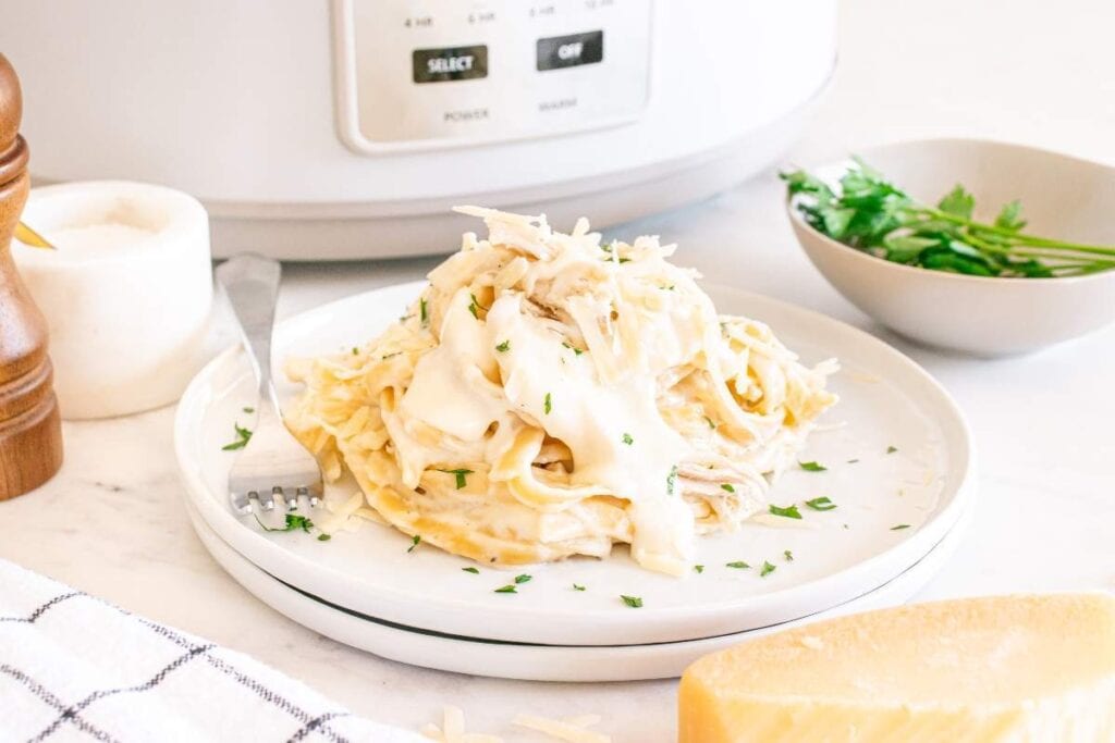 A plate of fettuccine Alfredo with shredded chicken, perfect for quick pasta recipes, is garnished with parsley and sits in front of a slow cooker, next to a bowl of fresh herbs and a wedge of cheese.