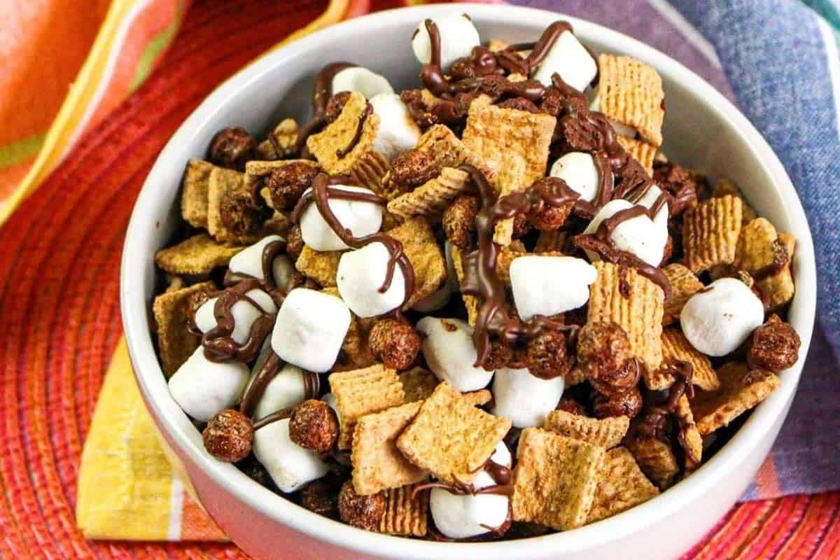 A bowl filled with cereal squares, chocolate cereal balls, mini marshmallows, and drizzled chocolate, placed on a colorful surface.