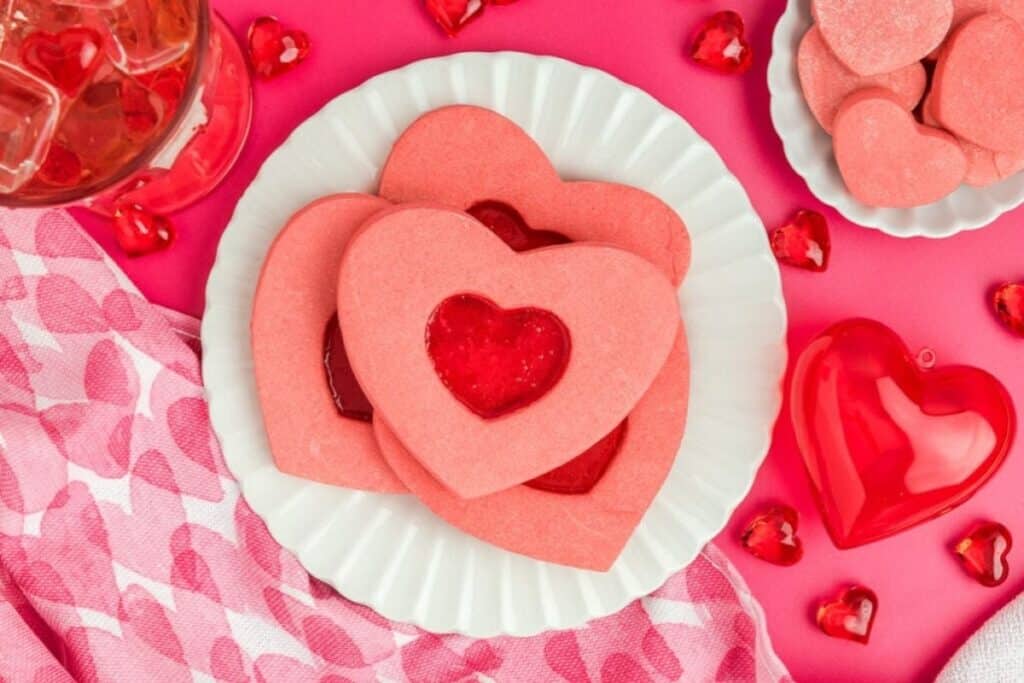 Heart-shaped pink cookies with a red jam center are arranged on a white plate, making perfect Valentine&rsquo;s desserts, surrounded by heart decorations on a pink background with a patterned napkin.