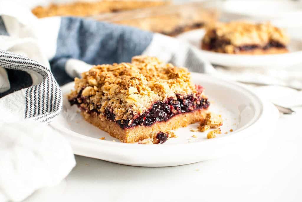 A square piece of berry crumble bar with a crumbly topping sits on a white plate, perfect for those who love Starbucks Copy Cat Recipes, with another piece and a striped napkin in the background.